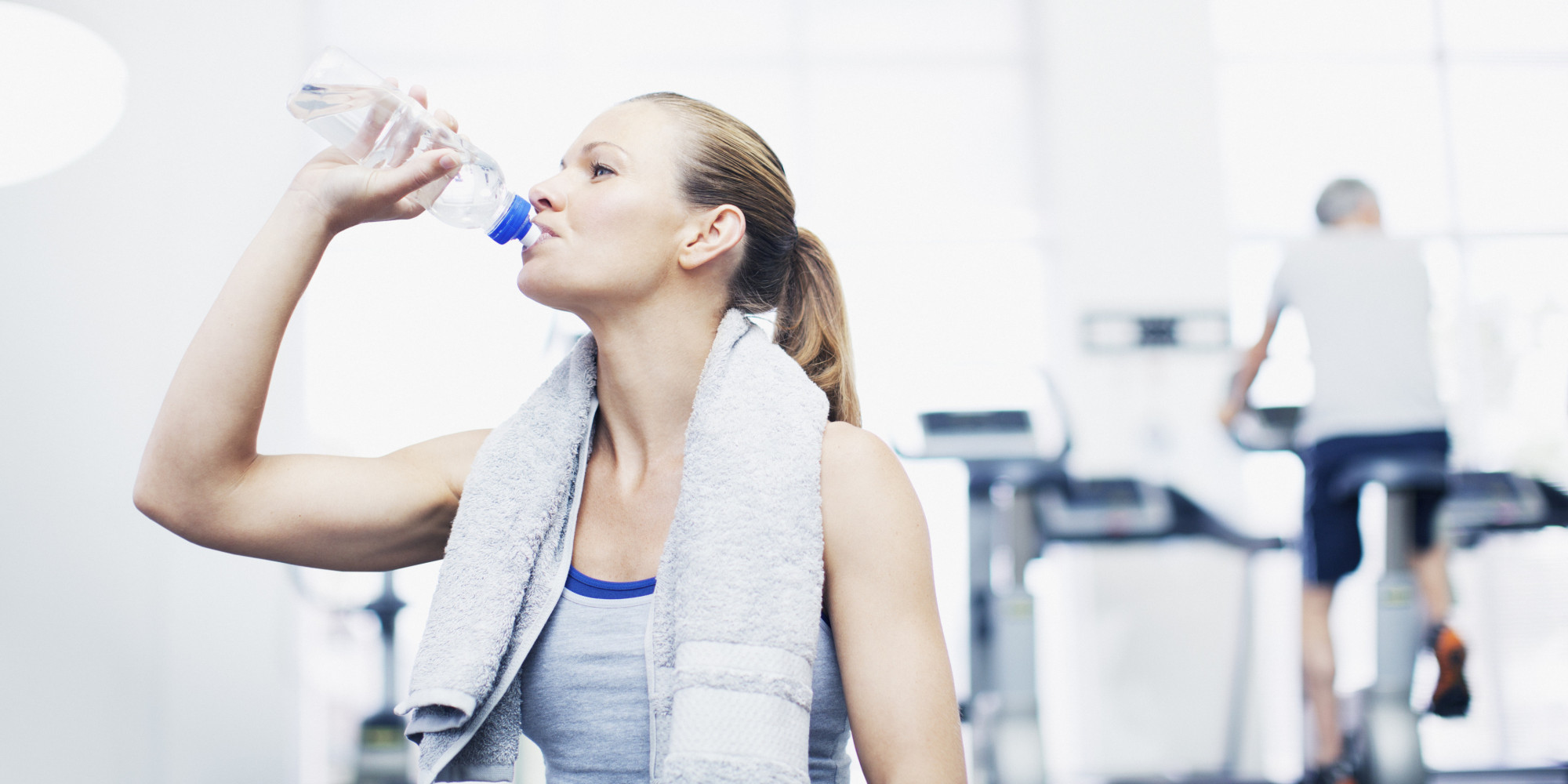Woman with towel around neck drinking water in gymnasium